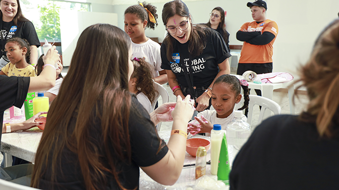 Adults and children smile as they make slime together at a long indoor table.