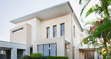 A modern two-story house with a light exterior and flat roof, surrounded by palm trees and tropical plants.