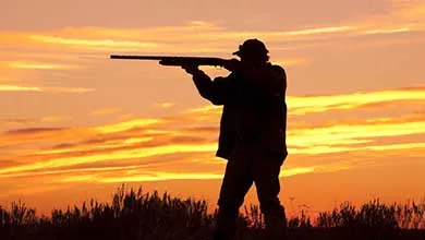 Silhouette of a hunter aiming a rifle in a field at sunset.