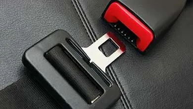Close-up of a car seatbelt tongue poised to enter a red and black buckle, set against a black leather seat.