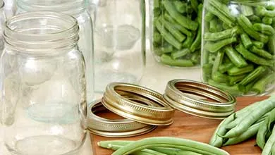 Empty and filled canning jars with green beans, fresh beans, and lids on a wooden cutting board.