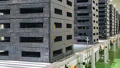 Stacks of dark gray plastic pallets move on a conveyor belt in a large warehouse.