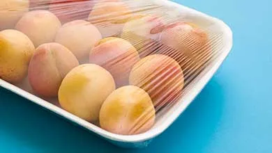 Close-up of fresh apricots in a white tray, covered with clear plastic wrap, on a bright blue background.