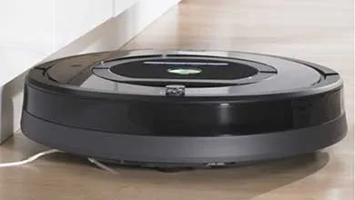 A black circular robotic vacuum with a green light on top sits on a wooden floor beside a white wall.