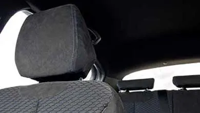 Close-up of a car’s front seat upholstered in dark gray fabric, featuring a suede-like headrest and a honeycomb pattern with blue stitching on the backrest. Rear seats are visible in the background.