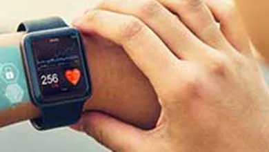 A smartwatch with a black band shows a heart rate of 256 and a red heart icon on its screen as a hand adjusts it on a wrist. Semi-transparent hexagonal icons with medical symbols—a cross, water droplet, and capsule—float above the skin against a blurred background.