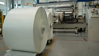 Large roll of white composite material positioned on a factory floor beside industrial manufacturing equipment.