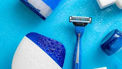 Men’s grooming products, including a blue-handled razor and blue and white toiletry containers, arranged on a bright blue surface with water droplets.